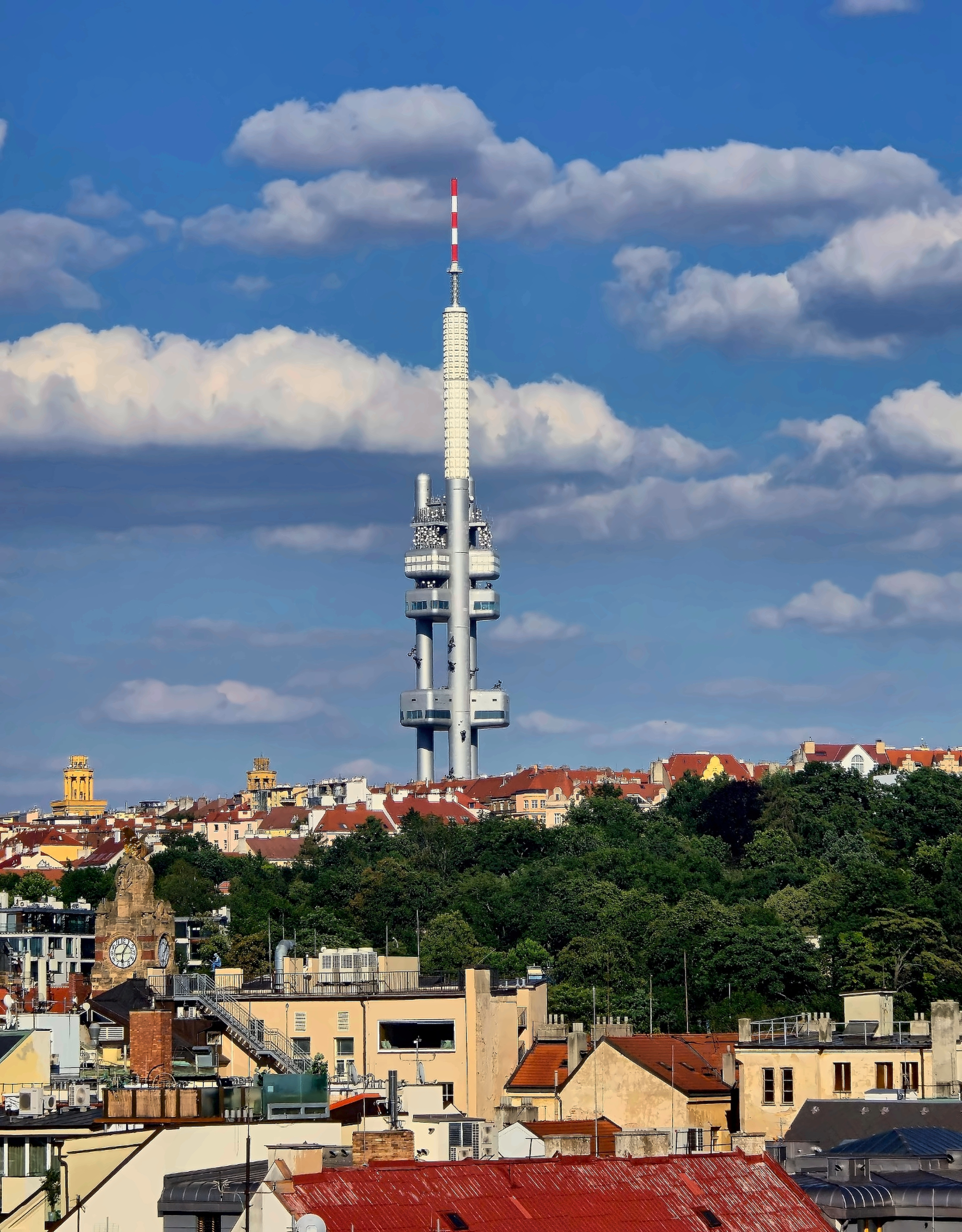 Žižkov Television Tower rises above Prague’s red-roofed buildings and leafy trees under a blue sky.