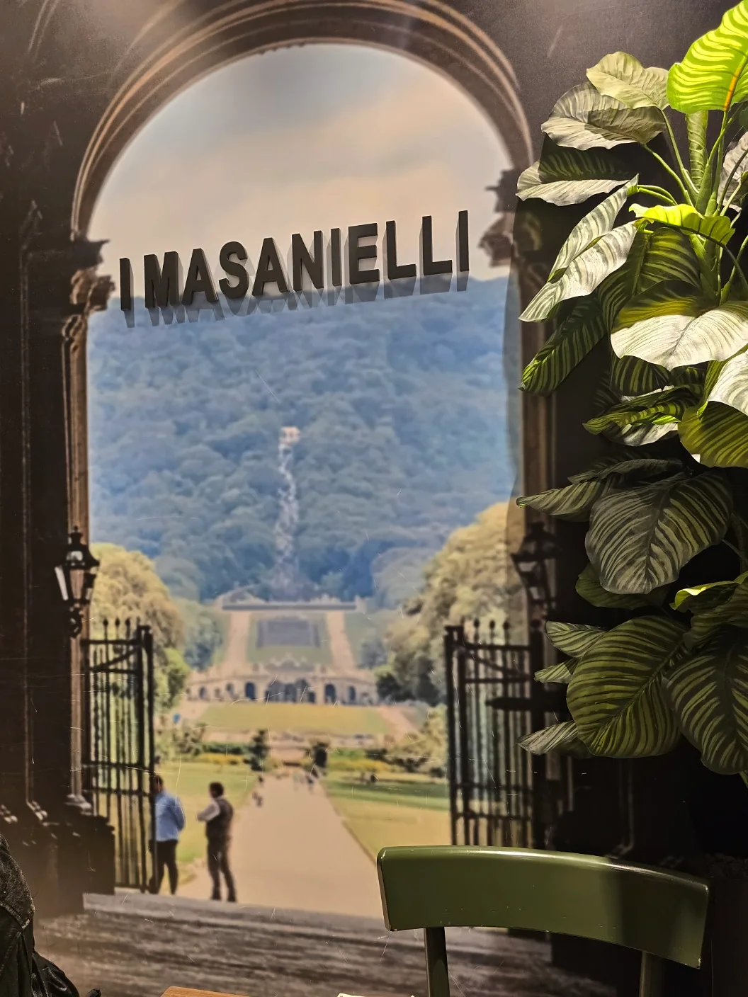 I Masanielli sign above a wall photograph of the Reggia park, beside a plant and green chair in Sasà Martucci's pizzeria.