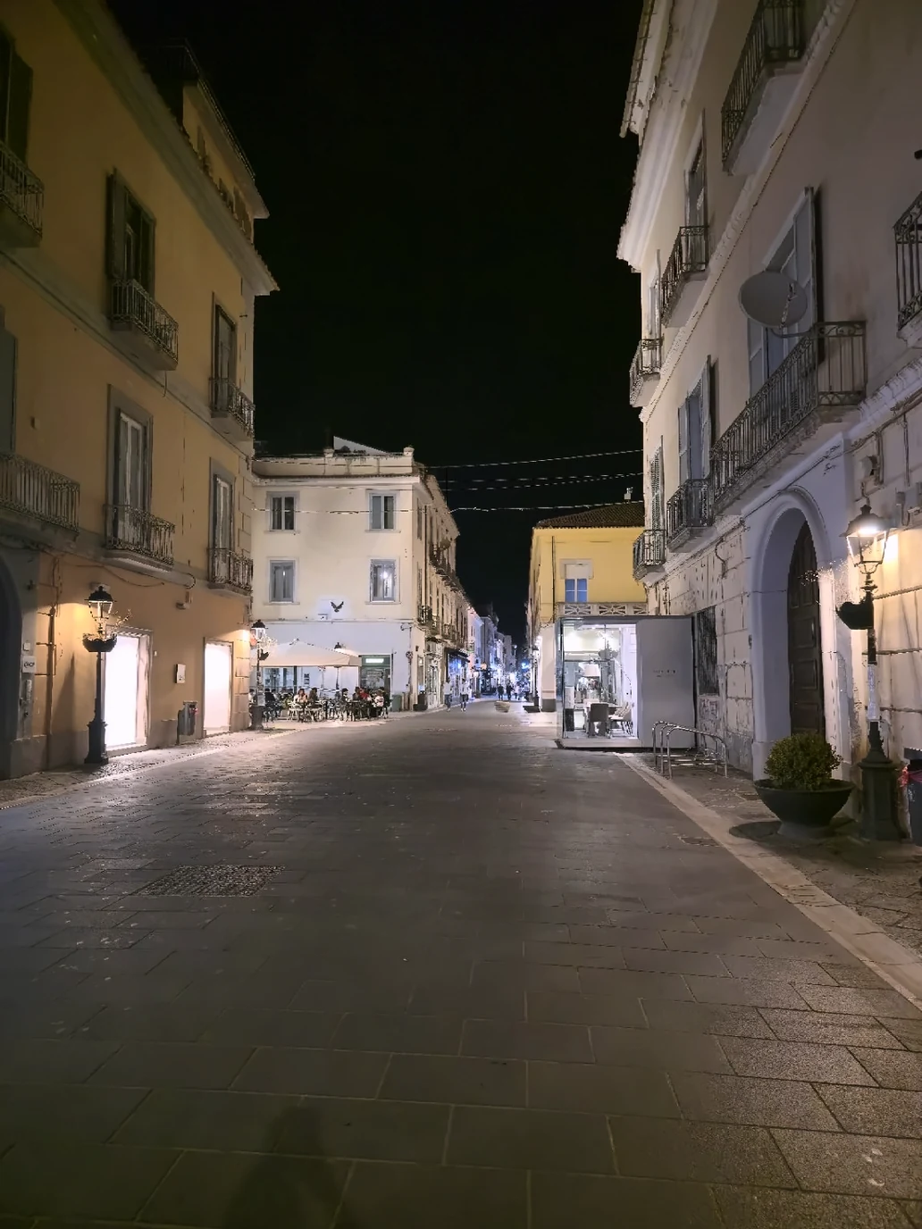 An evening street in central Caserta with illuminated shopfronts, balconies and a café terrace.