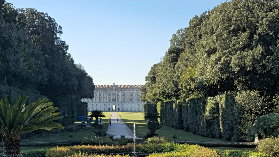 The Reggia di Caserta framed by trees and hedges, with a garden path and fountain in the foreground.