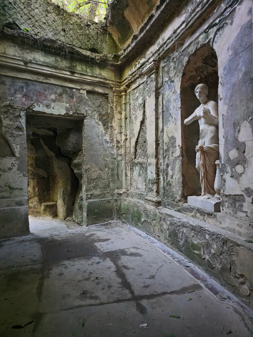Stone statue in a wall niche of the Caserta cryptoporticus, beside worn plaster, the open vault and a passage towards the garden.