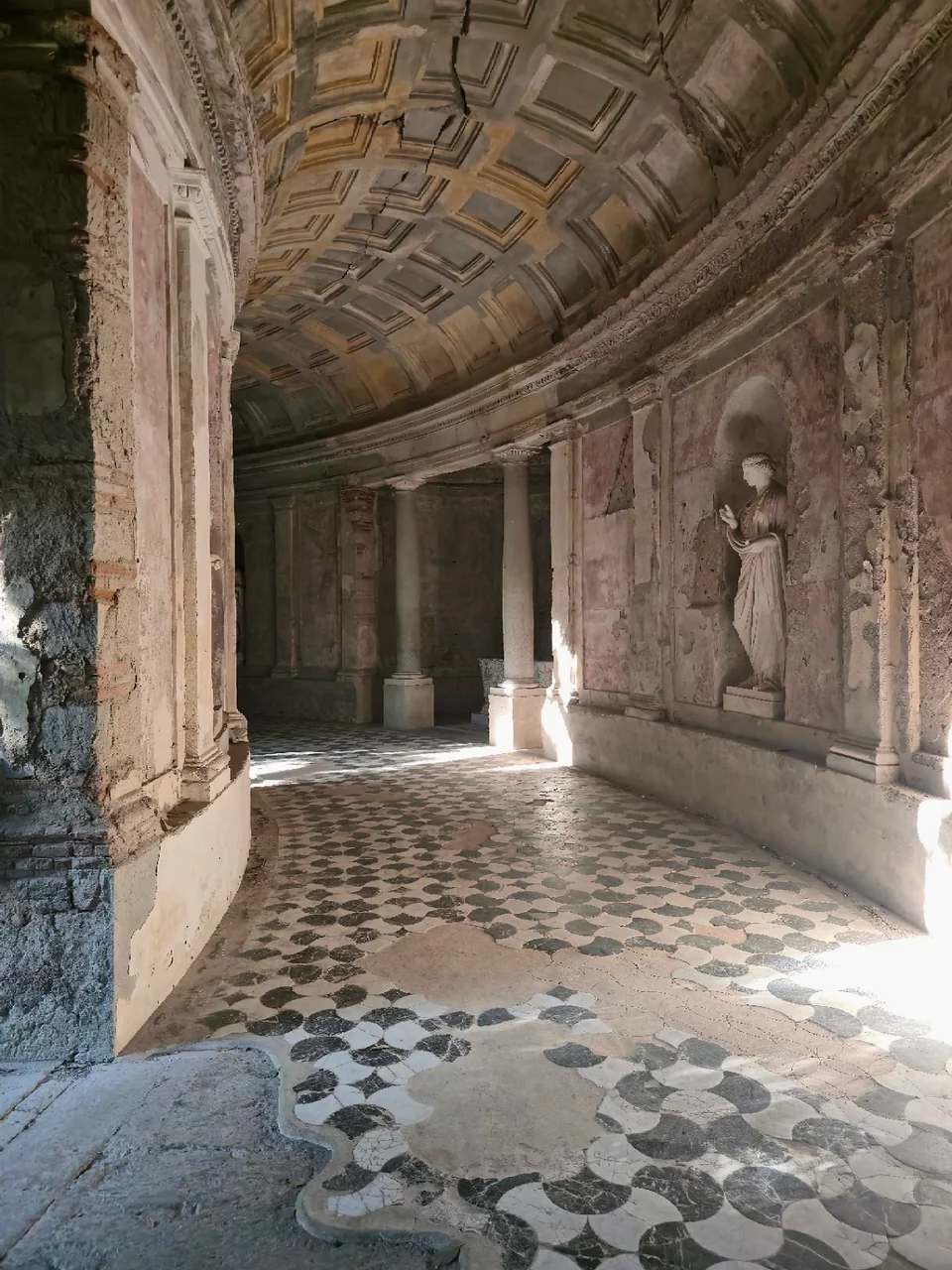 Curving gallery of the cryptoporticus in Caserta, with columns, a coffered vault, a statue in a niche and patterned stone paving.