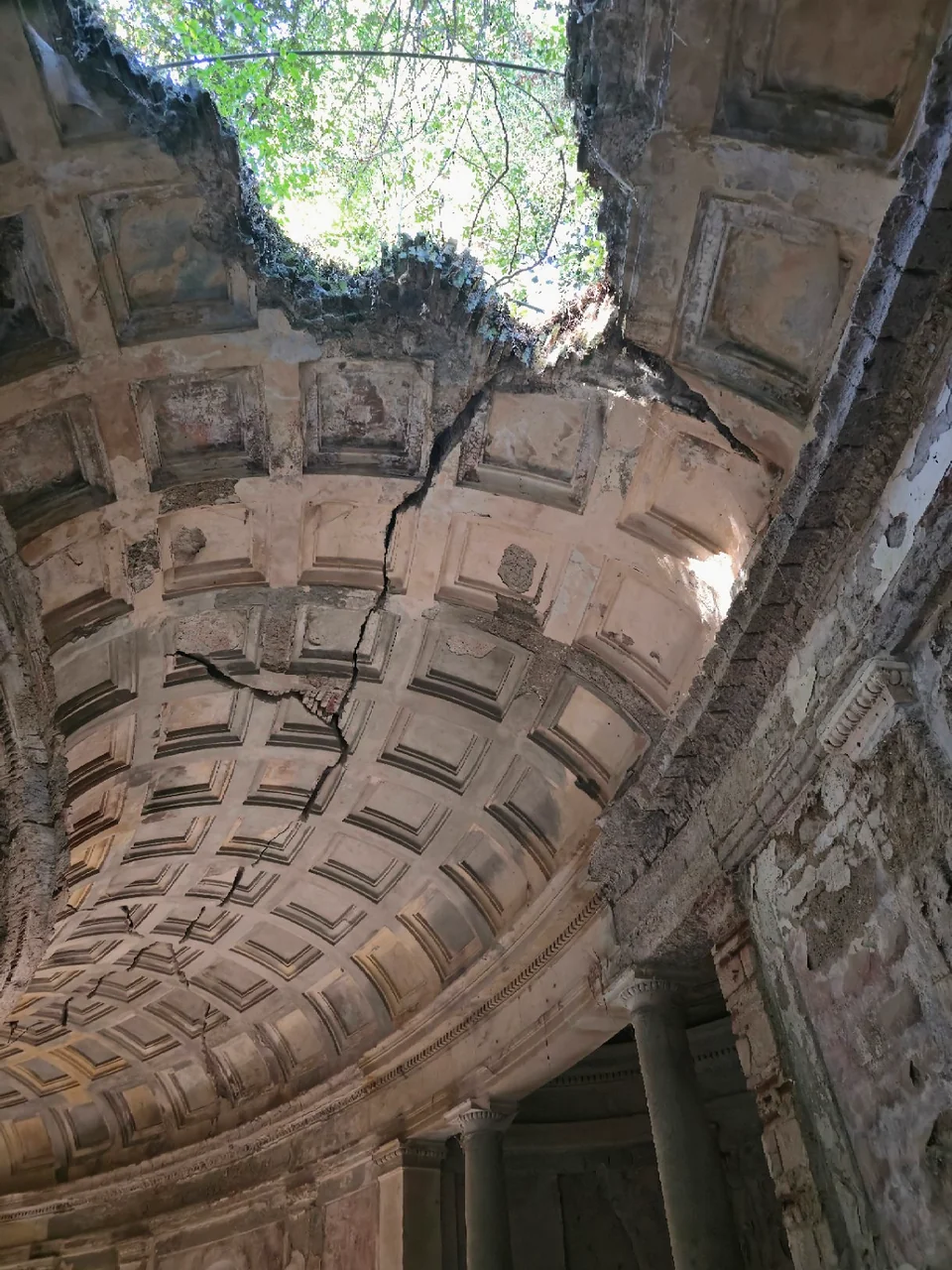 View towards an opening in the coffered vault of the cryptoporticus in Caserta, with cracks and branches visible above.