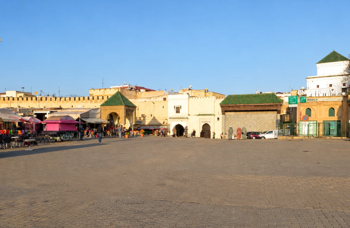 Market stalls and historic buildings line Place el Hedim in Meknes, with green-tiled roofs and few people in the square.