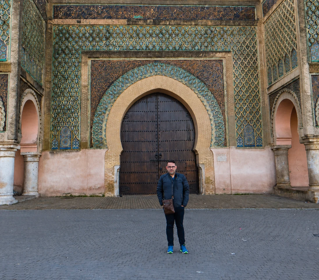 A man in a dark jacket stands before Bab Mansour’s ornate tiled gateway in Meknes.