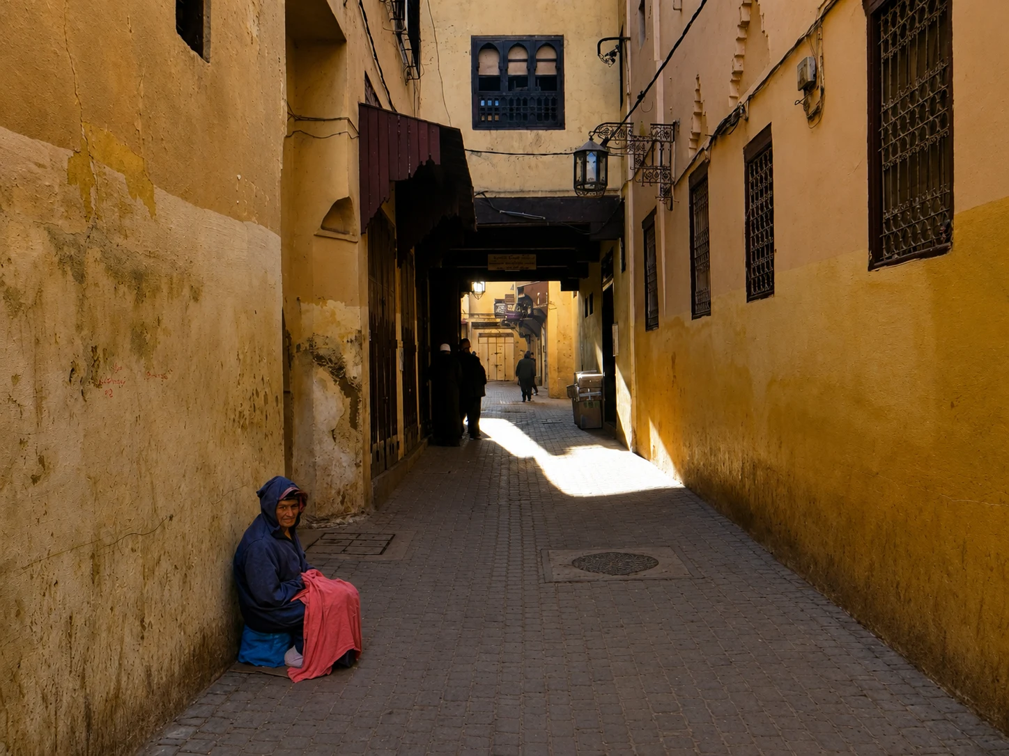 Seated person in a blue top and pink skirt beside a narrow, sunlit medina alley in Meknes.