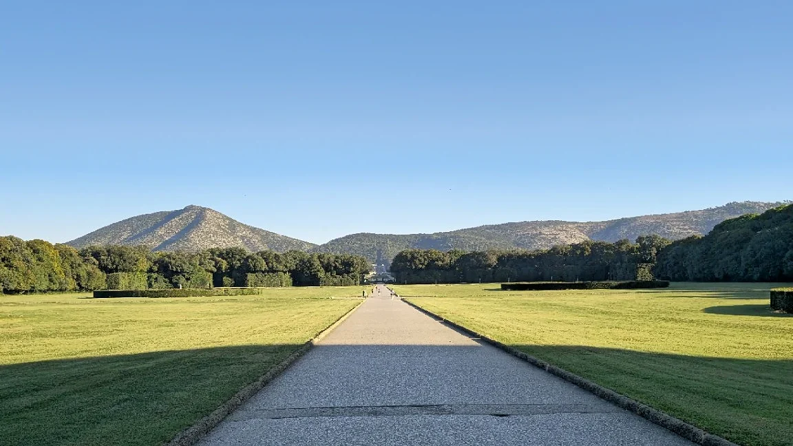 Long straight path through the park of the Reggia di Caserta towards the fountains and hills, with visitors in the distance.